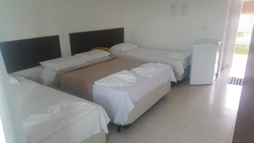 Quarto da Pousada Peruíbe Ametista. Uma cama de casal e uma cama de solteiro de cada lado. No fundo, no canto direito, um frigobar e a porta de entrada do quarto.