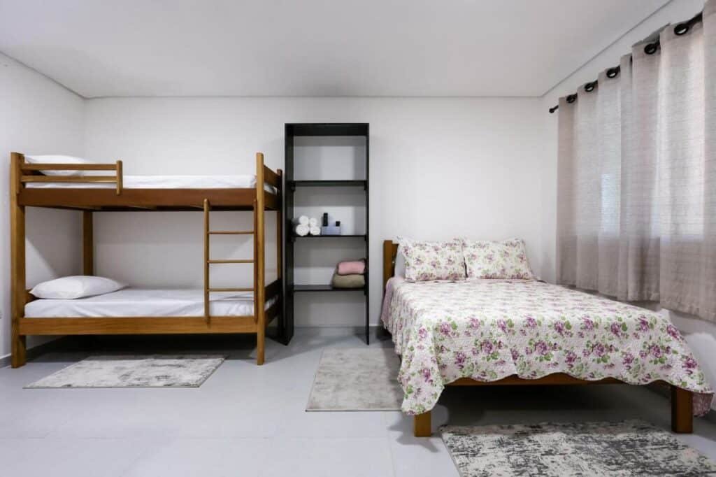 Quarto da Pousada Peruibiza. Uma cama de casal no lado direito com a janela do lado. No lado esquerdo uma cama beliche. Entre as camas uma estante com três prateleiras.