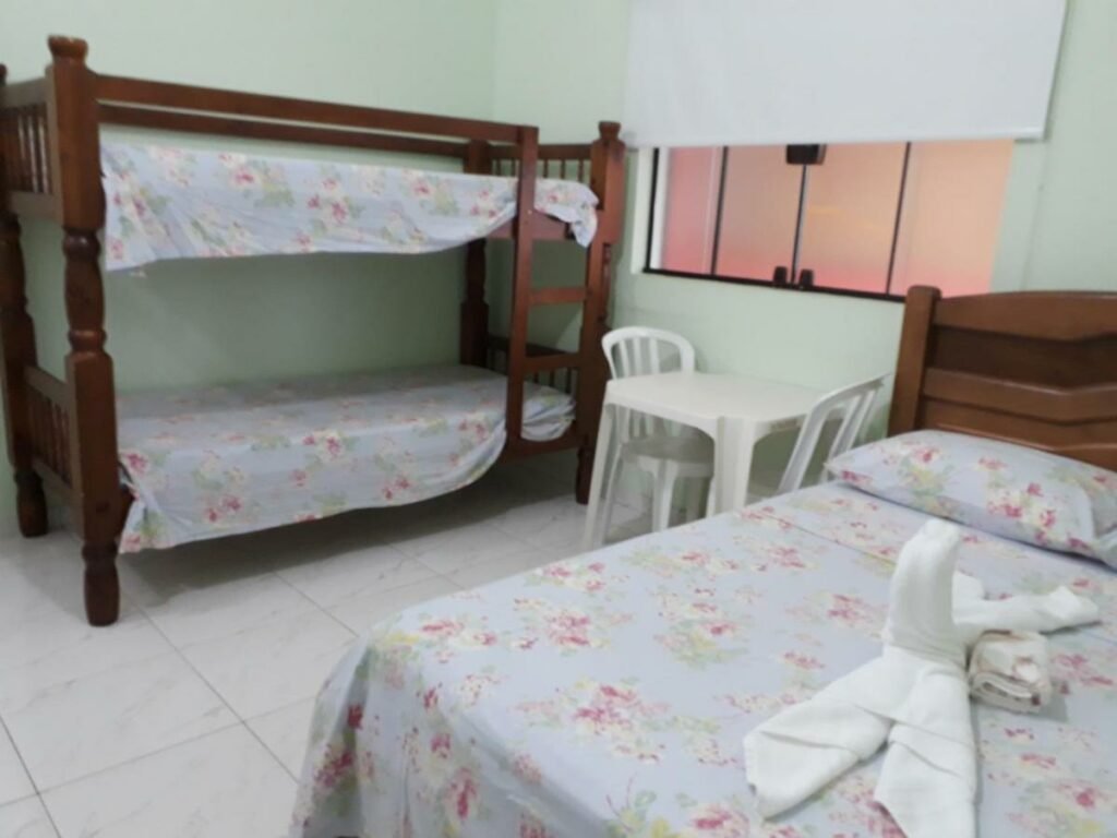 Quarto da Pousada Pesque Park. Uma cama de casal no lado direito, uma mesa de plástico no meio com uma janela. E no lado esquerdo uma cama beliche.
