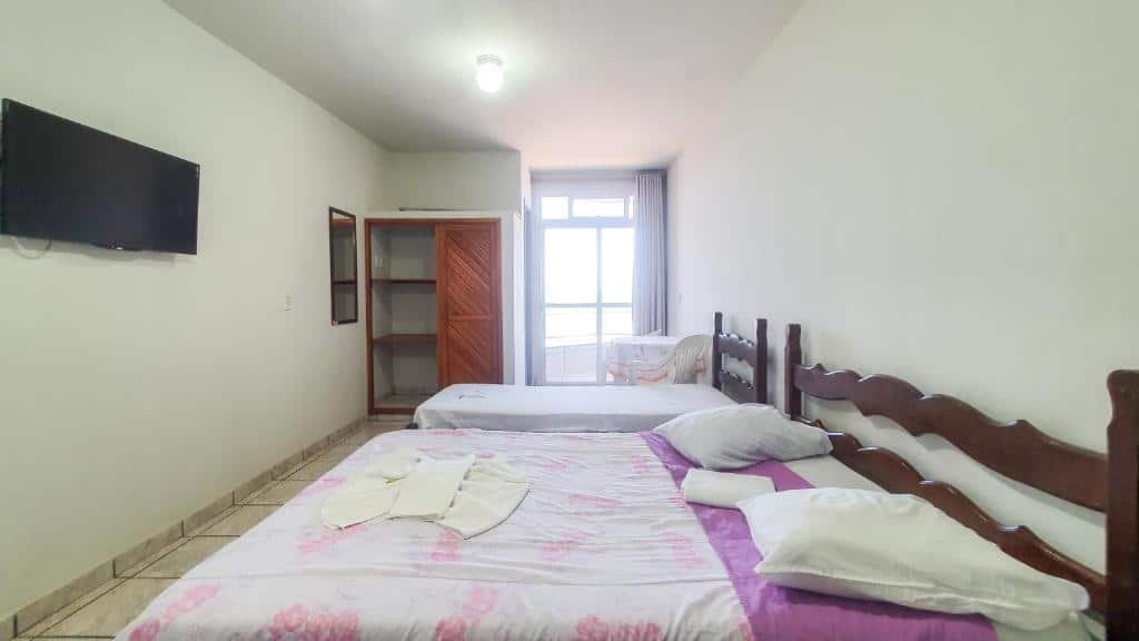 Quarto da POUSADA POLONINI. Uma cama de casal e uma cama de solteiro no lado direito, de frente uma televisão e um espelho. No fundo, no lado esquerdo um guarda-roupa. Do lado direito uma mesa com duas cadeiras e a porta da sacada.