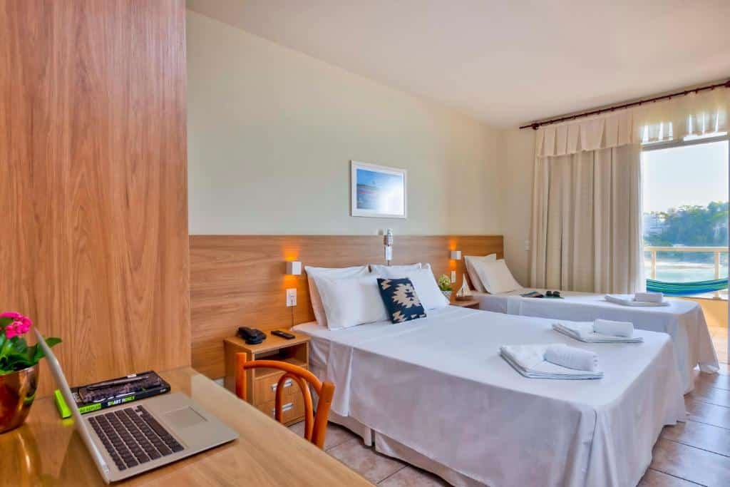 Quarto do Hotel Portal da Lua. Uma mesa de trabalho com um computador e um guarda-roupa no lado esquerdo. De frente, uma cama de casal e atrás um cama de solteiro, com cômodas entre elas. No fundo, atrás da cama de solteiro, a sacada do quarto, com uma rede e vista para o mar. Foto para ilustrar post de pousadas em Iriri (ES).