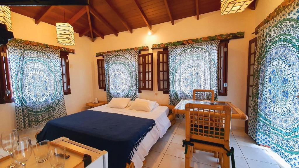 Quarto da Pousada Pura Vida Maresias com cama de casal, mesa de madeira e vidro com duas cadeiras do lado direito e a frente frigobar, o quarto possui quatro janelas de vidro com cortinas azuis.