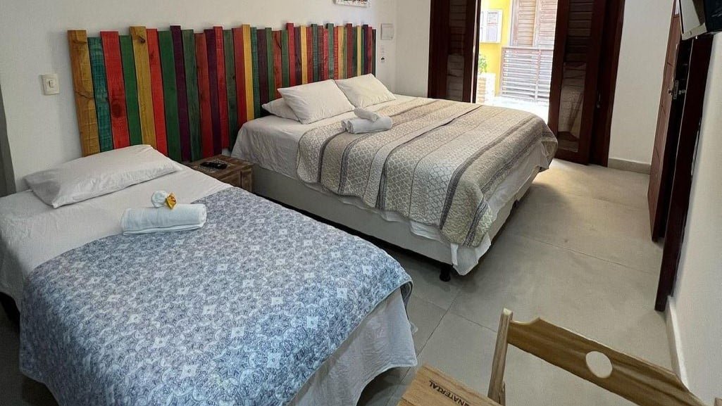 Quarto triplo da Pousada ResDelMar. Uma cama de solteiro está no canto esquerdo e uma cama de casal está no canto direito, ao lado da porta. Na parede em frente a elas existe uma tv e um pouco mais ao lado há uma mesa com cadeira.