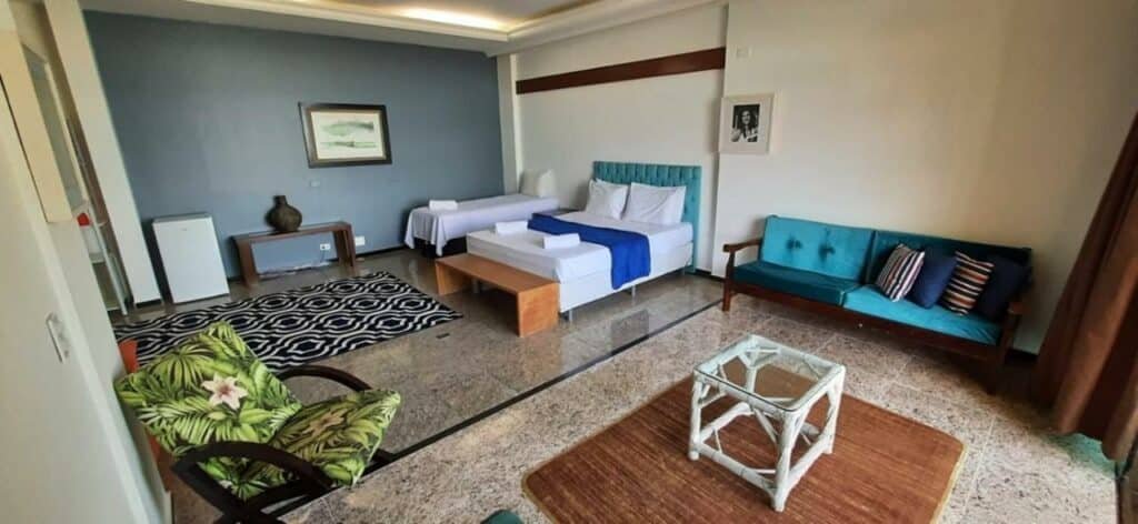 Quarto da Pousada Stand Up! Guarujá com uma ampla varanda, local com dois ambientes, um sendo uma pequena sala com sofá e poltrona, e no outro o quarto com a cama de casal e uma de solteiro, além do frigobar e itens de decoração