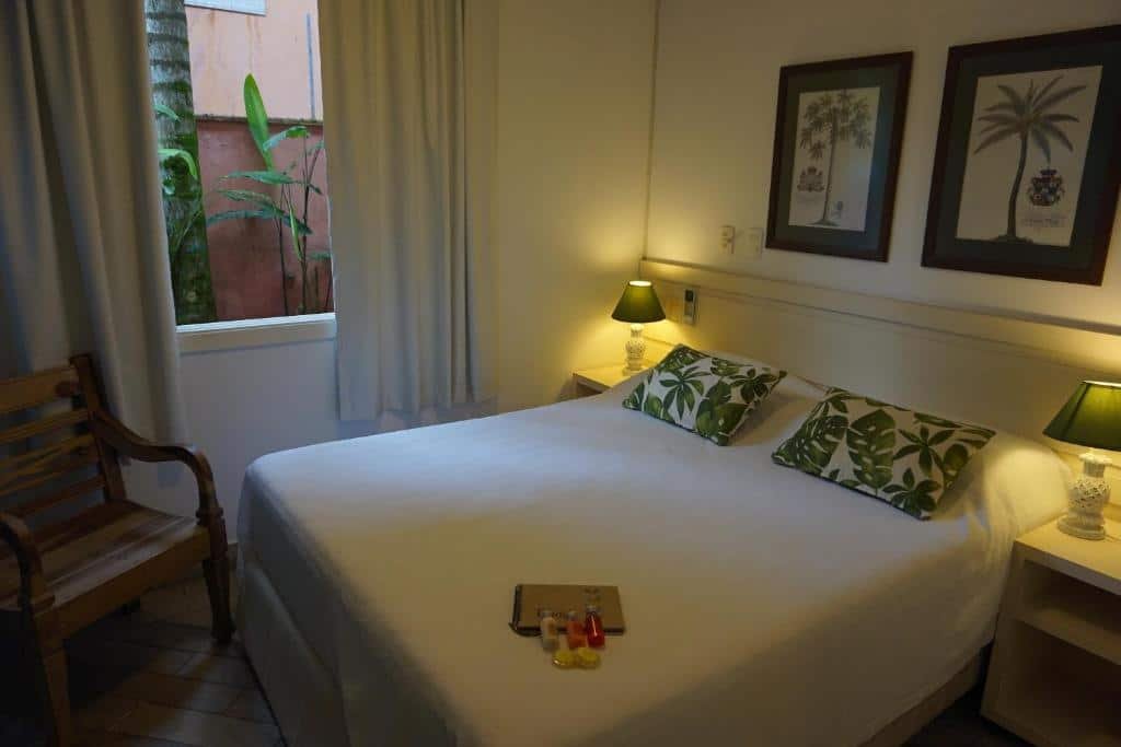 Quarto da Pousada Terra Juquehy com cama de casal, duas cômodas de ao lado da cama com luminárias, cadeira de madeira do lado esquerdo perto da janela com cortinas brancas.