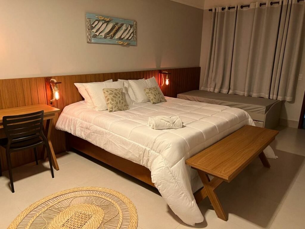 Quarto na Pousada Timoneiro, com uma cama de casal, uma mesa e uma cadeira, dois pequenos abajures, uma cama de solteiro, uma janela com cortina, para representar pousadas em Vila Velha