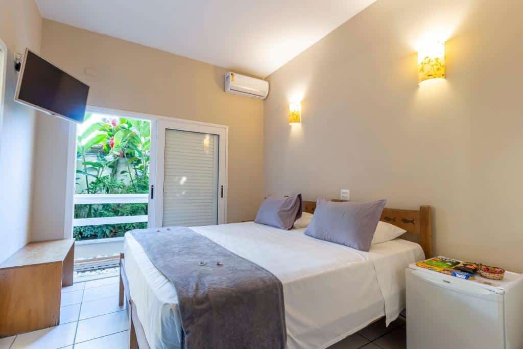 Quarto da Pousada Toca da Praia com cama de casal ao meio, frigobar do lado direito, TV em frente a cama presa na parede, cômoda de madeira abaixo e, do lado esquerdo, há portas de madeira que dá acesso à varanda.