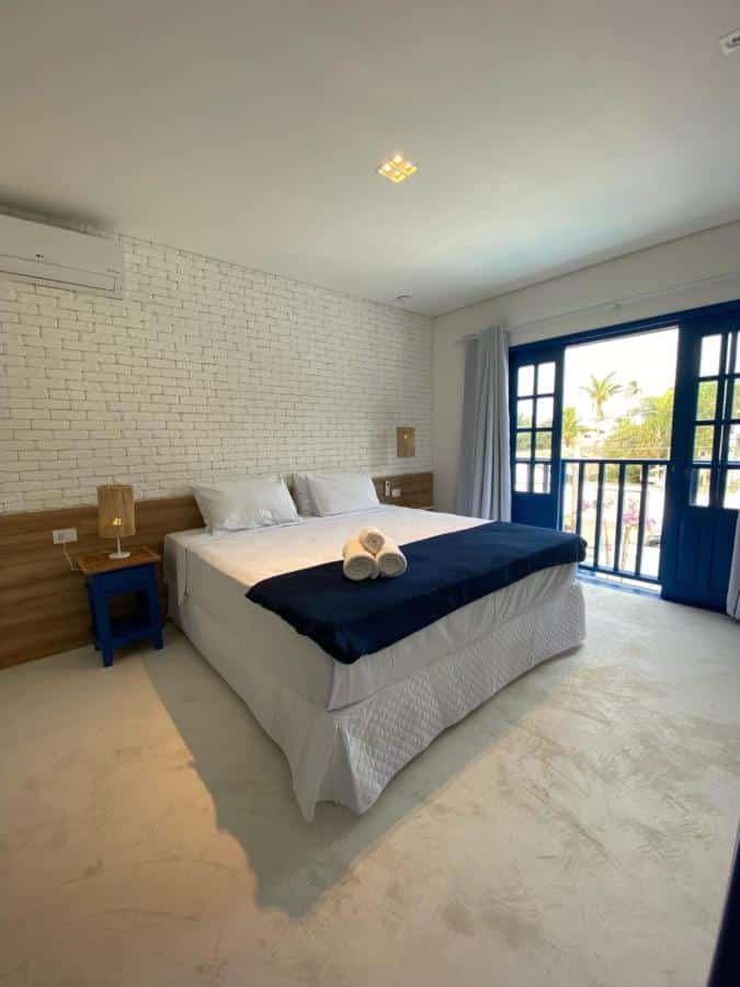 Quarto da Pousada Vila Santô com tudo decorado em branco e azul, uma cama de casal com travesseiros e toalhas, uma ampla sacada, um ar-condicionado e dois abajures