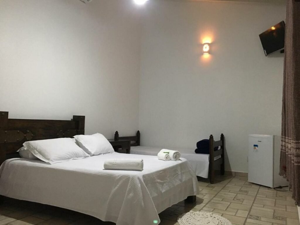 Quarto da Villa de Cananea. Uma cama de casal está no meio do quarto e, ao seu lado, há uma cama de solteiro encostada na parede. No meio delas é possíver ver uma mesinha. Também encostado na parede e em frente a cama de solteiro está um mini frigobar. Mais acima está a tv. 