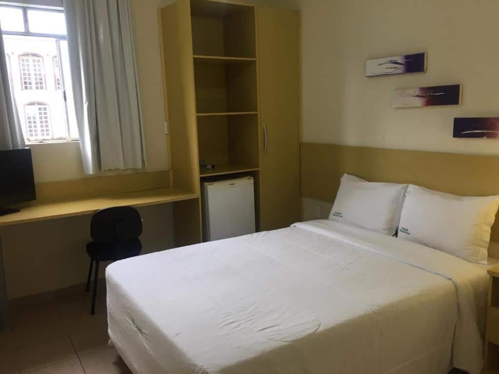 quatro da Pousada Vista da Serra em Ouro Branco com uma cama de casal, uma mesa de trabalho com uma cadeira preta e uma televisão em cima, e uma armário de MDF no canto com um frigobar embaixo