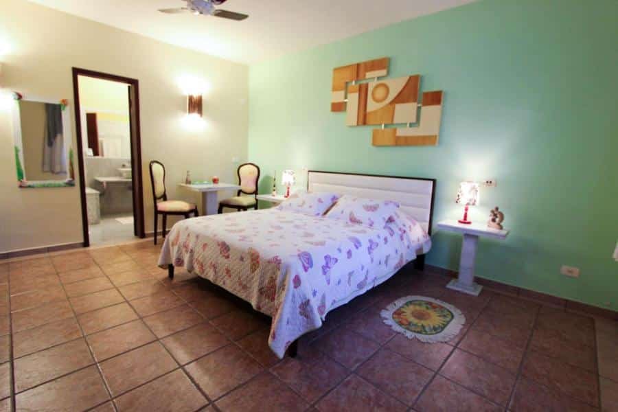 Quarto Pousada Vivenda do Flamboyant, local amplo com uma cama de casal, um banheiro, um espelho, ao lado da cama tem abajures, e uma mesa com duas cadeiras