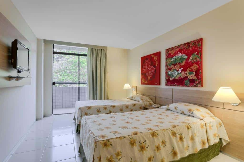 Quarto do Praiabella Hotel com uma pequena varanda com cortinas, duas camas de solteiro com uma cabeceira que tem abajures, sob as camas há dois quadros com pinturas vermelhas e, de frente para as camas, há uma televisão