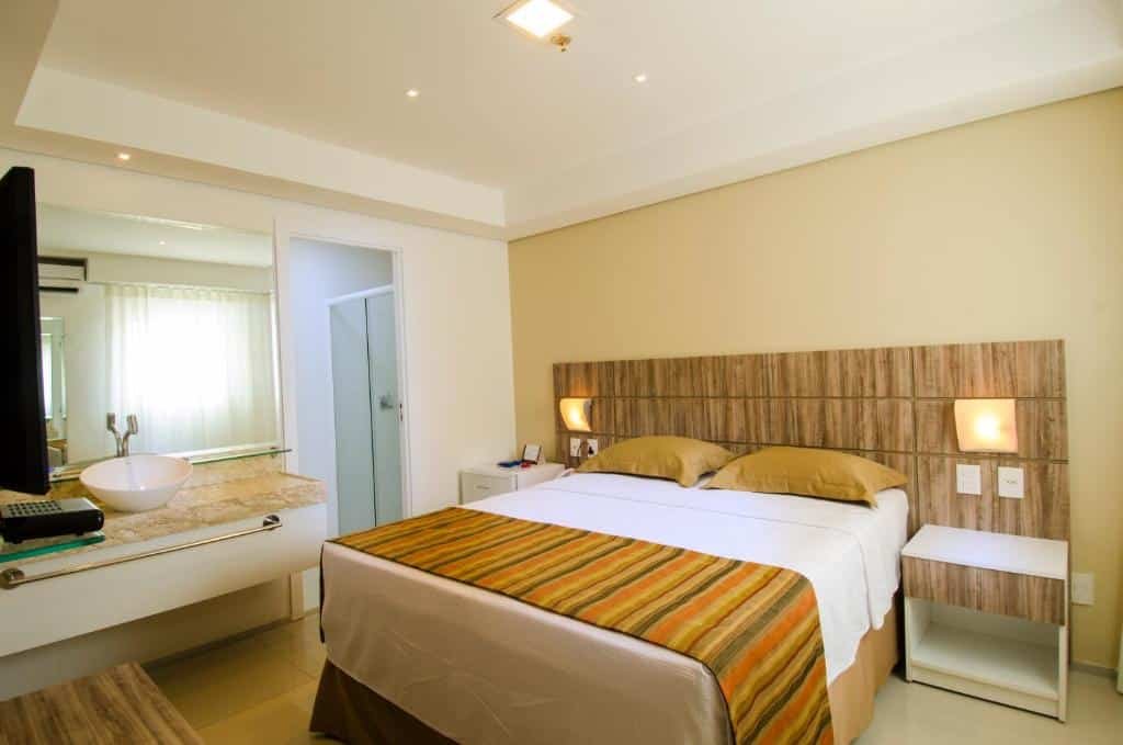 Quarto do Profissionalle Hotel São Luís com uma cama de casal, uma cabeceira de madeira com luminárias, de frente para a cama há uma televisão e um móvel de madeira, e do lado esquerdo do quarto há uma pia e a entrada para o banheiro, para representar hotéis em São Luís do Maranhão