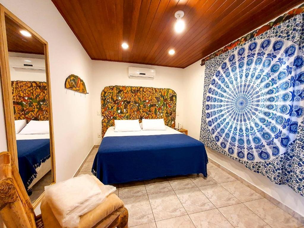 Quarto da Pousada Pura Vida Maresia com cama de casal no centro do quarto, cortina azul do lado direito e do lado esquerdo um espelho grande.