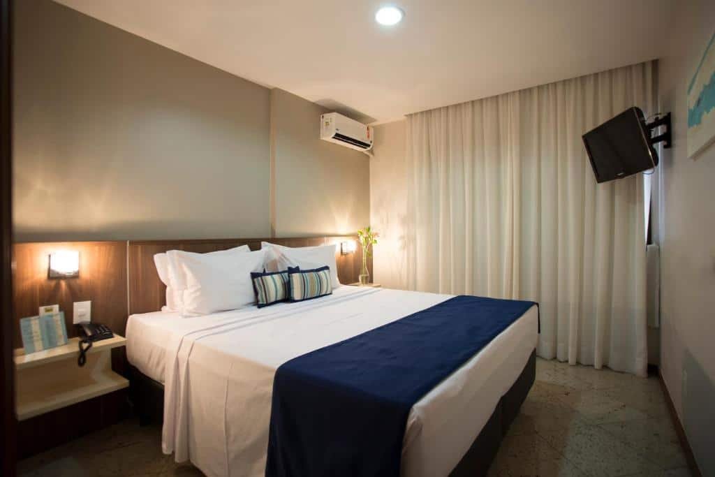Quarto no Quality Suites Vila Velha com uma cama de casal, uma televisão, duas mesinhas de cabeceira, uma varanda com cortinas e um ar-condicionado, para representar pousadas em Vila Velha