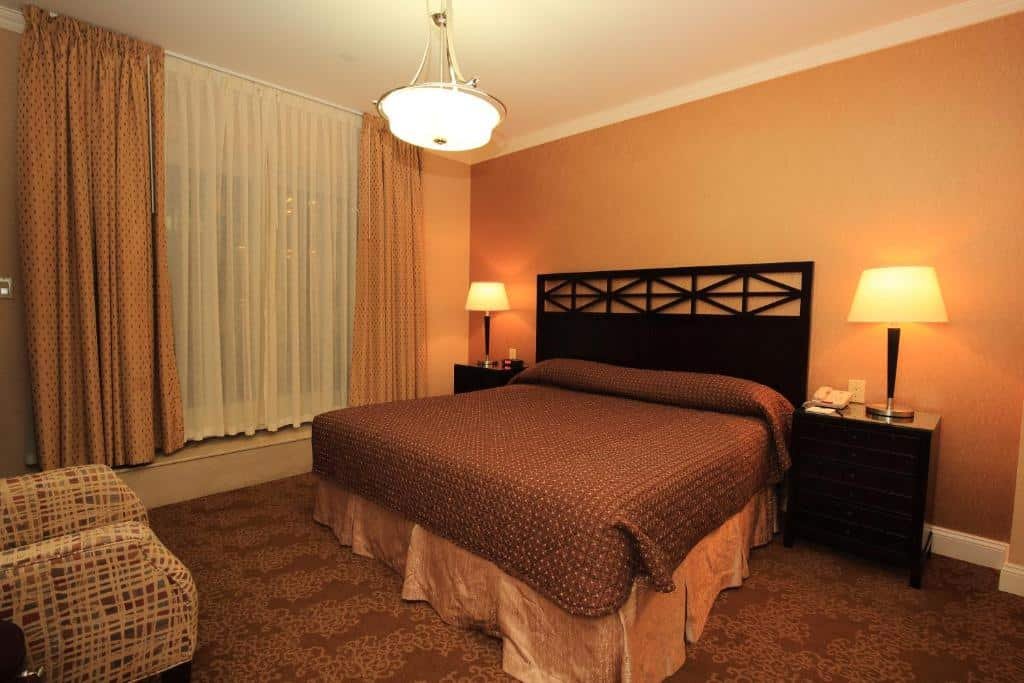 quarto do Radio City Apartments com uma cama de casal ao centro do espaço, duas cômodas, uma de cada lado da cama, com dois abajures acesos e uma poltrona listrada disposta em frente a cama, no canto inferior esquerdo da imagem.