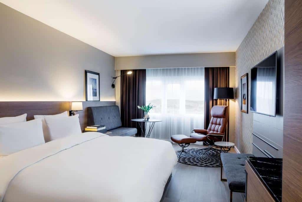 Quarto do Radisson Blu Plaza Hotel, Oslo com cama de casal, sofá cinza do lado esquerdo perto da janela e uma poltrona. Já a TV fica em frente da cama.