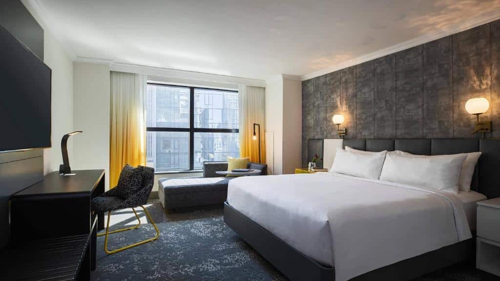 quarto do Renaissance New York Times Square Hotel com uma cama de casal à direit, um sofá estilo divã no lado esquerdo da cama, embaixo da janela ampla e uma mesa preta com luminiária em frente a cama.