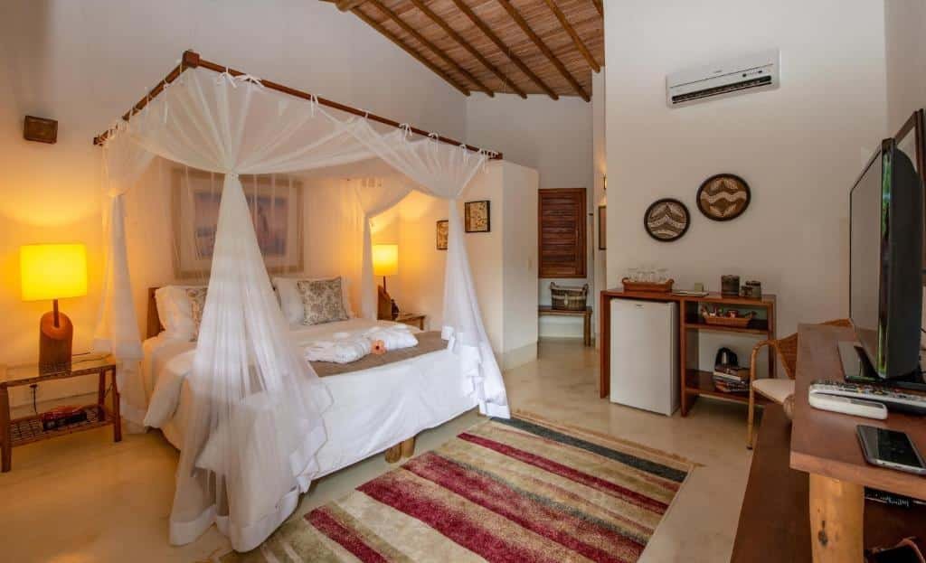 Quarto do Hotel e Resort Villas de Trancoso com cama de casal do lado esquerdo, com duas cômodas ao lado com luminária, do lado esquerdo da cama frigobar e em frente a cama cômoda com TV.