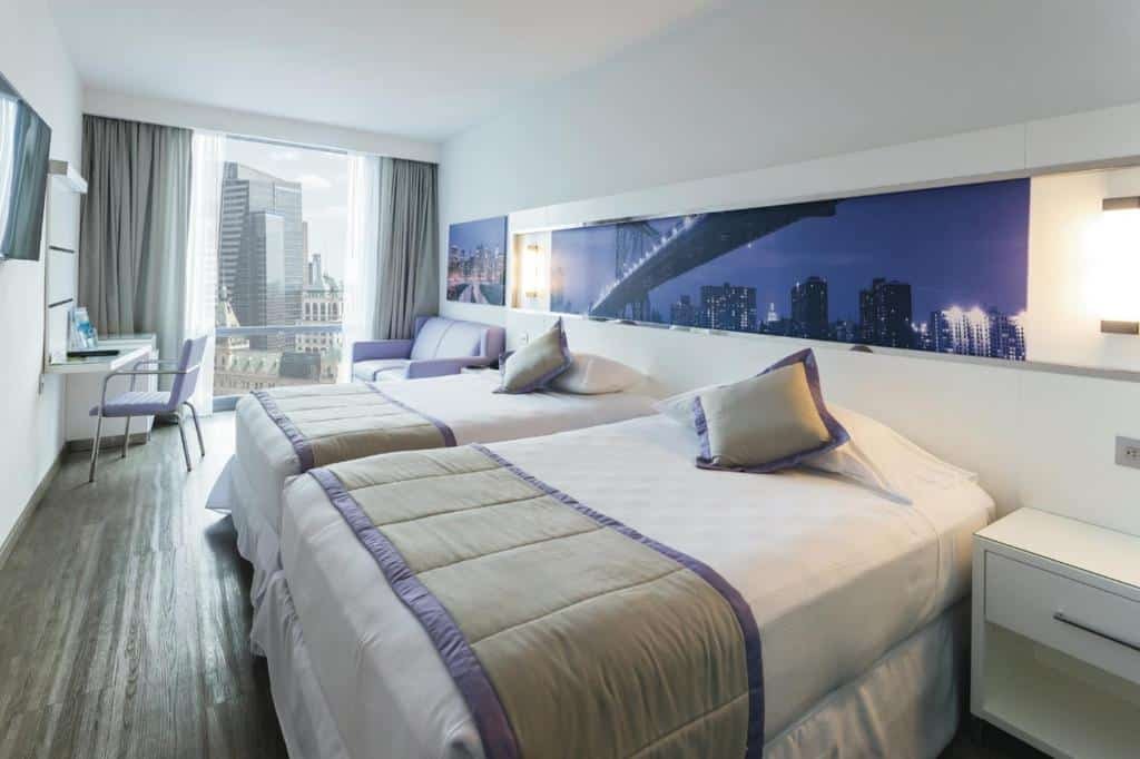 quarto do Riu Plaza New York Times Square com duas camas de casal à direita da imagem, uma ampla janela ao fundo do quarto com vista para os prédio de Nova York e uma pequena mesa com cadeira, em frente a janela, no lado oposto às camas.