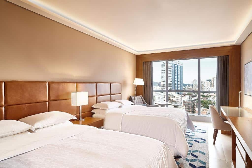 Quarto do Sheraton Santos Hotel com uma grande janela, uma cama de solteiro e uma de casal com roupas de cama brancas, uma mesa ampla de escritório com uma cadeira, o chão é de madeira e tem um tapete cinza com detalhes em azul