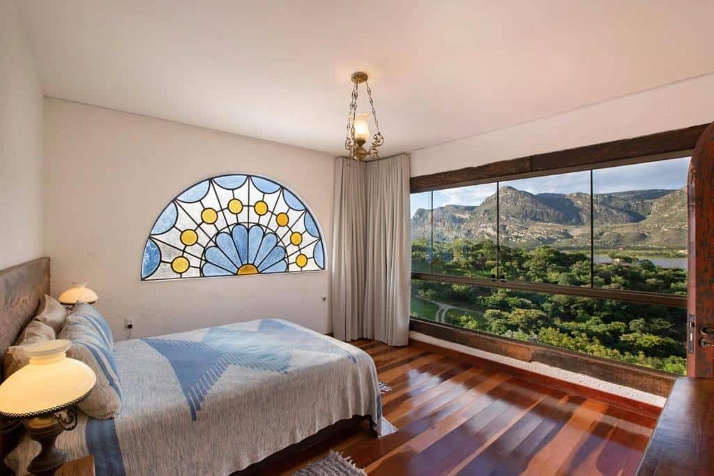 quarto da pousada Sítio Jatoba na Lapinha da Serra com uma cama de casal à esquerda, um vitral de flores azul, branca e amarela na parede ao fundo e uma ampla janela de vidro com vista para a Serra do Cipó mostrando várias árvores, um lago e lindas montanhas.