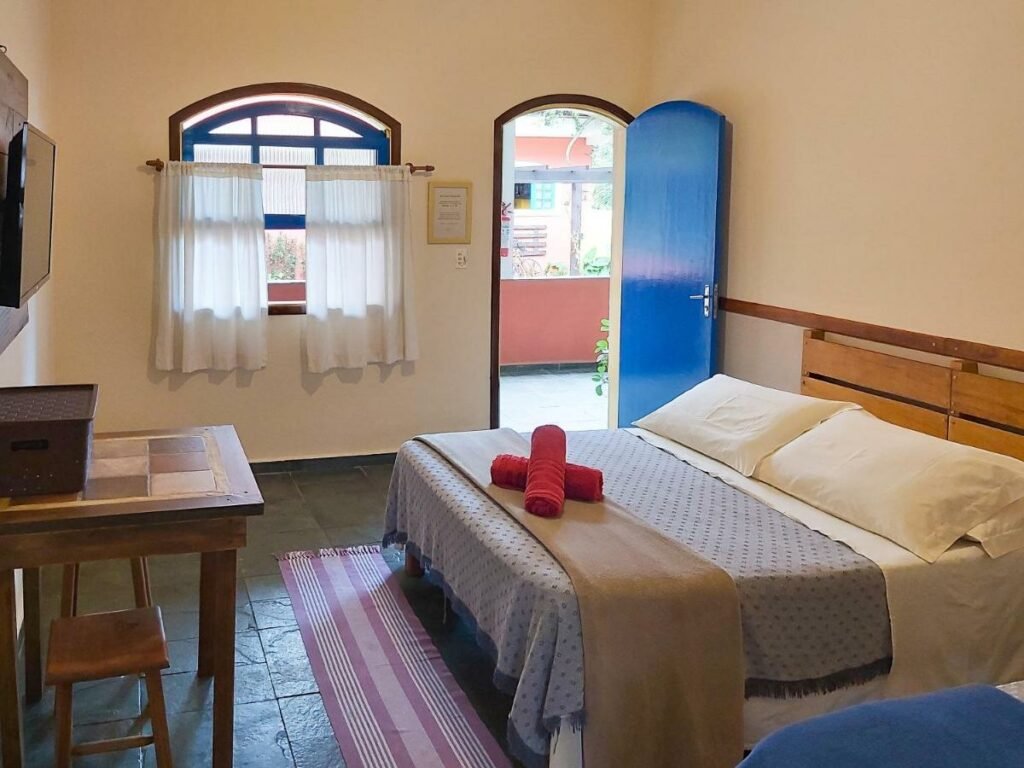 Quarto no Sollar dos Gerânios. Uma cama de casal no lado direito e atrás a porta do quarto com a janela. De frente para a cama uma mesa e a televisão.