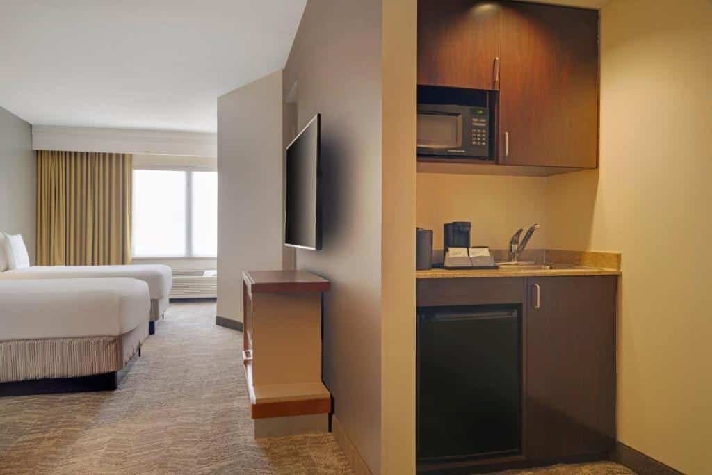 Imagem de um quarto no SpringHill Suites Newark Airport. No centro do quarto, há duas camas queen-size com lençóis brancos e travesseiros macios. Ao lado das camas, há mesas de cabeceira com luminárias de leitura. Na parede oposta às camas, há uma TV de tela plana montada. Ao lado direito da imagem, há uma área de estar com um sofá-cama e uma mesa de centro. Próximo à janela, encontra-se uma escrivaninha com uma cadeira. O quarto é iluminado de forma suave e possui uma atmosfera acolhedora. O SpringHill Suites Newark Airport é um dos hotéis perto do aeroporto de Newark.