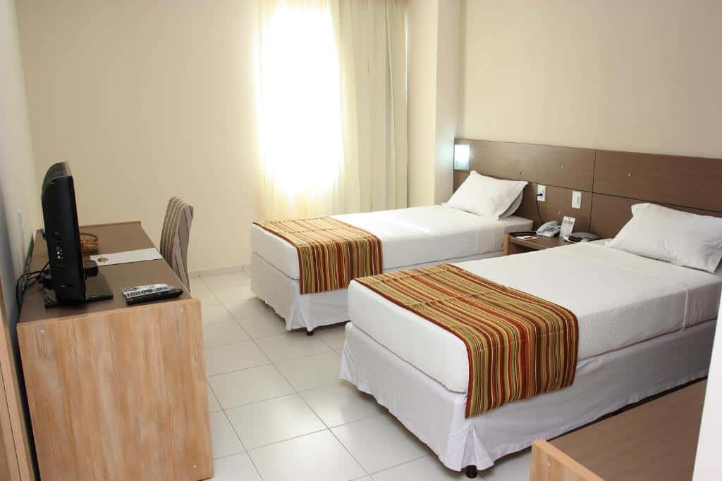 Quarto no Stop Way Hotel São Luís com duas camas de solteiro, uma janela com cortinas e, de frente para as camas, uma mesa de escritório com um cadeira e a televisão sob ela, para representar hotéis em São Luís do Maranhão
