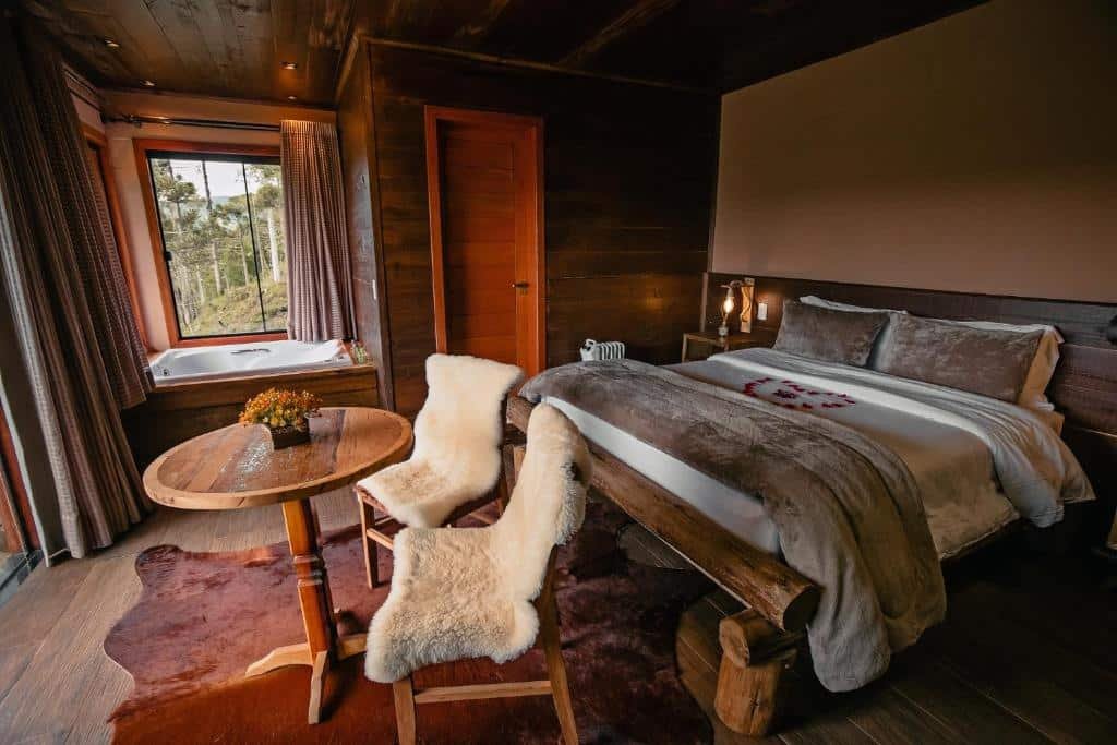 Quarto do Sunset Serrano Chalés em Urubici, com cama grande ao centro, mesinhas e luminária de ambos os lados, com cobertores pesados em cima, bem em frente, há uma mesa de tampo de madeira e duas cadeiras com encosto e assento como se fosse uma cobertinha, há ainda uma banheira abaixo de uma janela com vista da natureza.