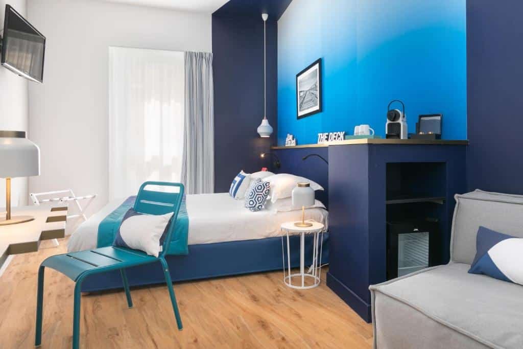 Quarto do The Deck Hotel by Happyculture. A cama está centralizada no quarto, no fundo há uma janela grande, em frente há uma mesa com abajour, cadeira e uma TV apoiada na parede. Na parte da frente do quarto há um pequeno balcão com uma cafeteira em cima e uma estrutura em baixo onde é encaixado um pequeno frigobar, ao lado há um sofá. Imagem para ilustrar o post hotéis em Nice.
