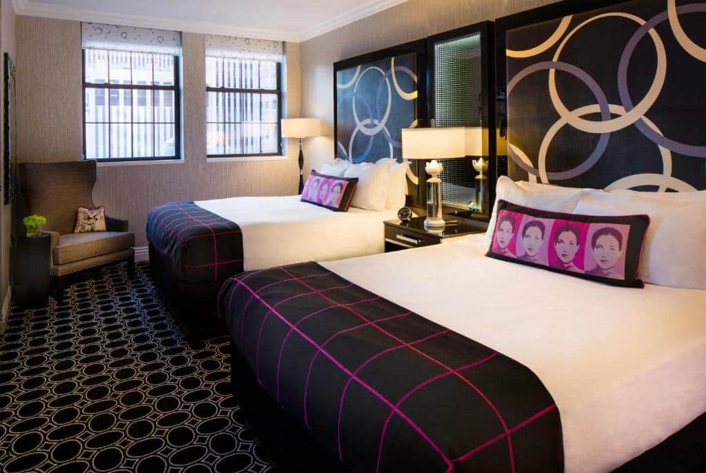 quarto do The Muse New York perto da Times Square com duas camas de casal à direita da imagem, com almofadas retangulares com rostos de mulheres em tons rosas impressos nas capas. No canto do quarto, lado esquerdo, ao fundo, há uma poltrona cinza abaixo das janelas quadriculadas.