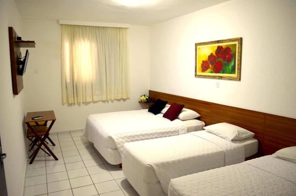Quarto com três camas, uma de casal e as outras de solteiro. Há também uma janela com cortina, mesinha e uma televisão.