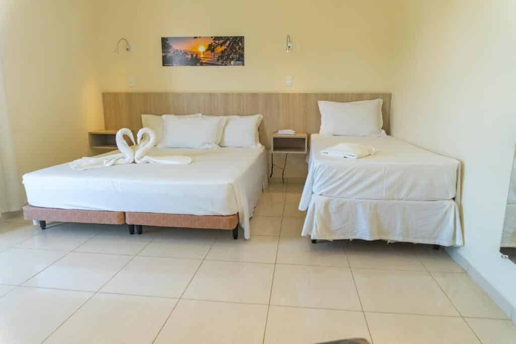 Quarto no Ubatuba Praia Hotel, uma cama de casal, uma de solteiro, um espelho e um quadro, para representar pousadas na Praia do Tenório