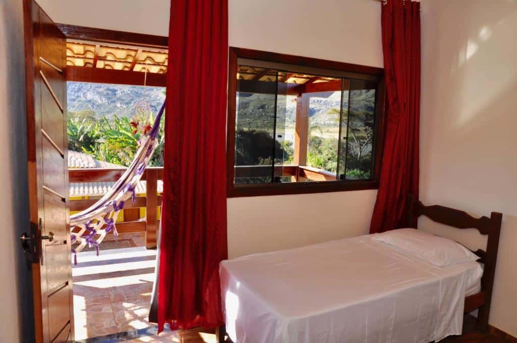 quarto da Vila dos Sonhos, uma das pousadas na Lapinha da Serra. A suíte mostra uma varanda coberta com vista para as montanhas e uma rede quadriculada, branca e roxa, pendurada do lado de fora. Dentro é possível ver um uma cama de solteiro logo abaixo da janela, em frente a porta