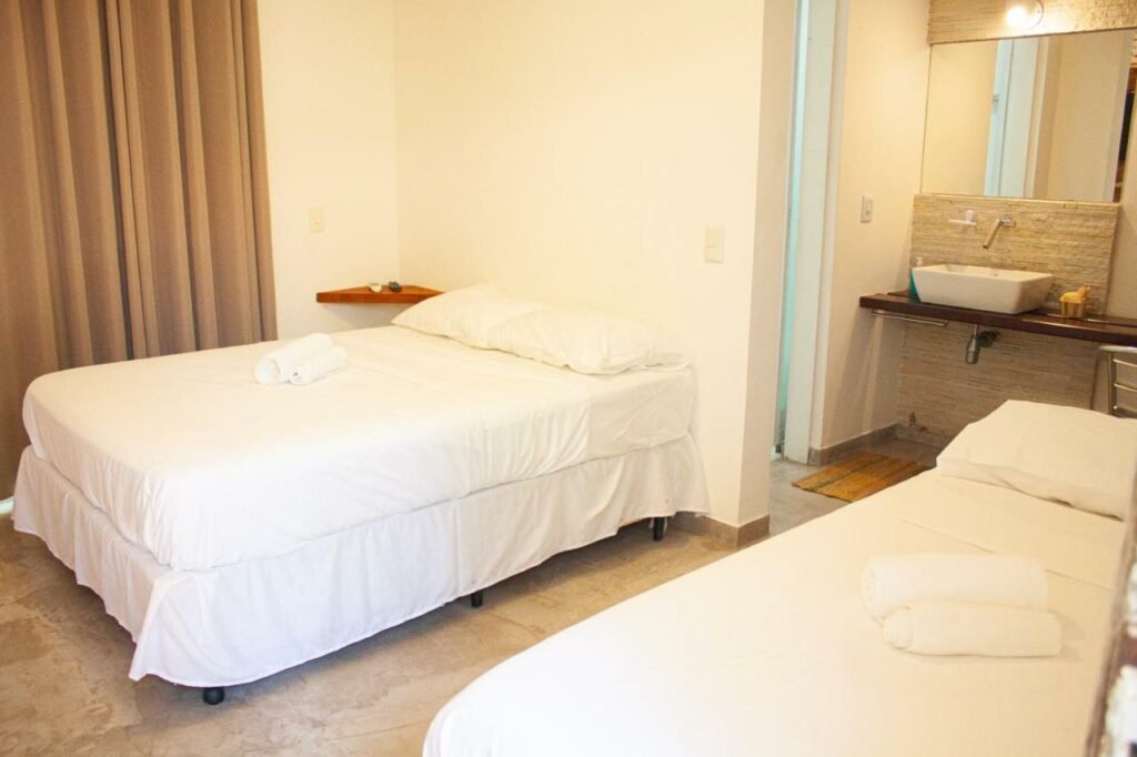 Quarto na Vila Jabutis com uma cama de casal e uma de solteiro, além de um banheiro e uma janela com cortinas