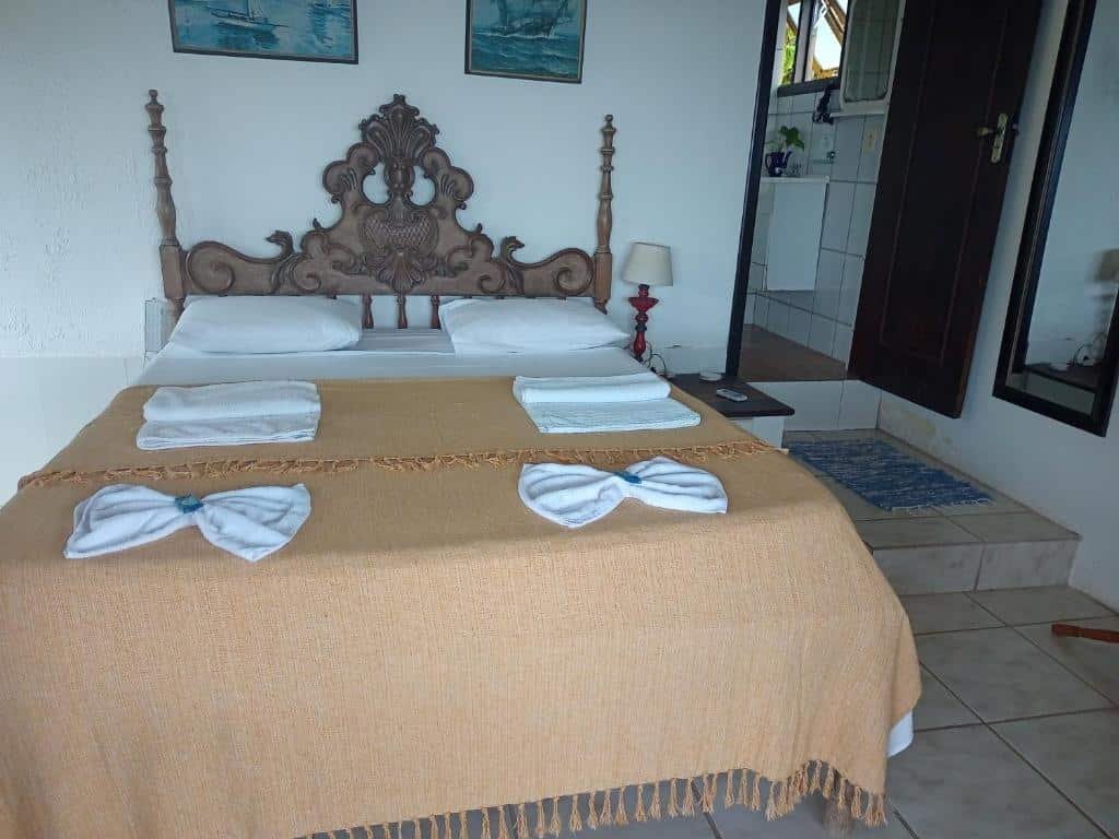 Quarto do Vila Lia com cama de casal, com toalhas em cima, cômoda ao lado da cama com luminária e ao fundo porta que dá acesso ao banheiro.