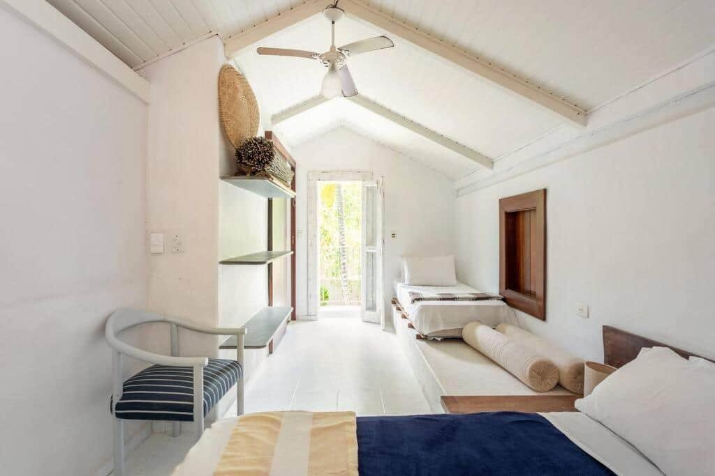 Quarto do Vila dos Tangarás com cama de solteiro em frente a imagem, cadeira de madeira do lado esquerdo e ao fundo outra cama de solteiro, com cômoda de madeira a frente.