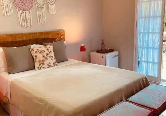 Quarto do Village Toque Toque com cama de casal, uma luminária acesa e frigobar ao lado, cama com travesseiros e almofadas. Representa airbnb na praia de Calhetas