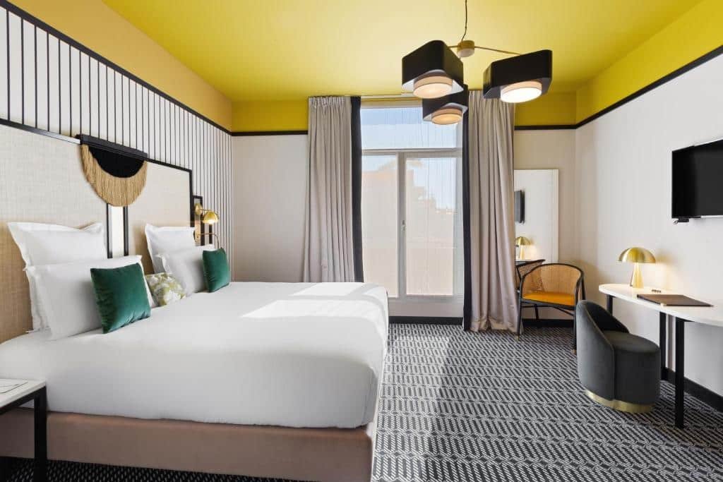 Quarto do Best Western Premier Hotel Roosevelt. A cama está encostada na parede do lado esquerdo do quarto, em frente a cama há uma mesa, uma pequena poltrona e uma TV a cima pendurada na parede. No fundo do quarto há uma porta de vidro que oferece acesso a varanda, um espelho e uma cadeira. Imagem para ilustrar o post hotéis em Nice.