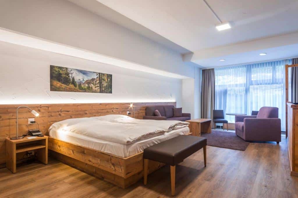 Quarto do Zermatt Budget Room com cama de casal do lado esquerdo da imagem no centro do quarto com uma comoda em cada lado com luminária no pé da cama um banco estofado e do lado esquerdo da cama ao fundo uma pequena área de estar com um sofá uma mesa de centro e uma poltrona. Representa onde ficar em Zermatt.