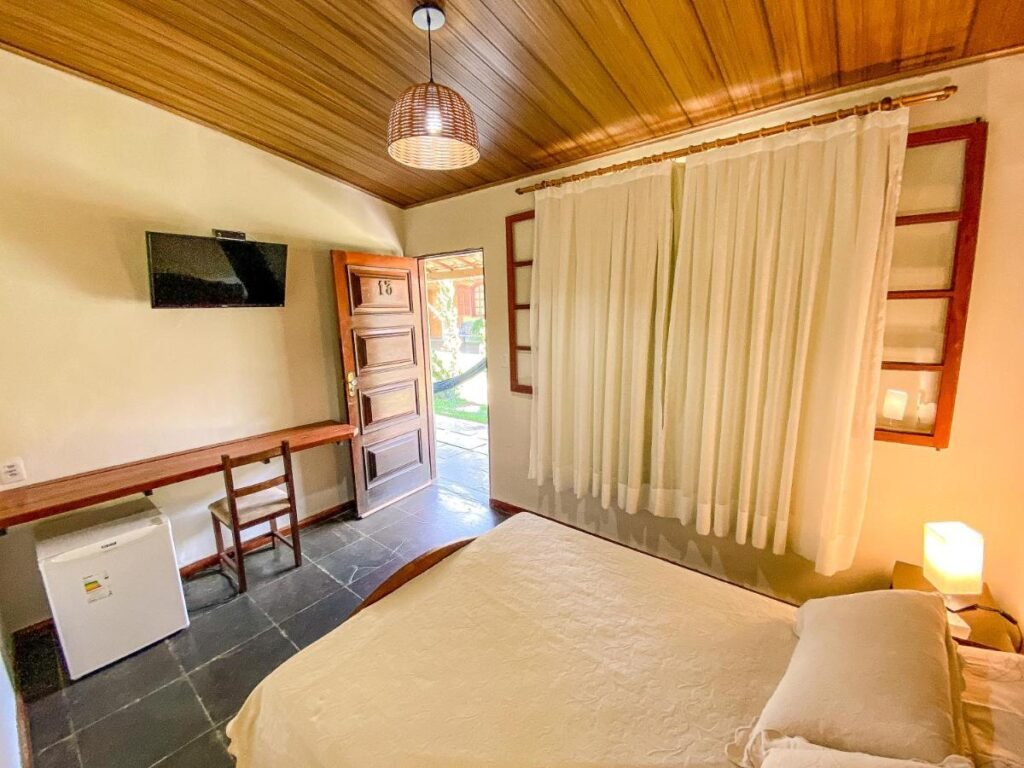 Um quarto aconchegante do Quatro Estações Pesqueiro e Hotel Fazenda com uma cama de casal, TV montada na parede, geladeira pequena, escrivaninha e cadeira de madeira, janelas com cortinas e teto de madeira.