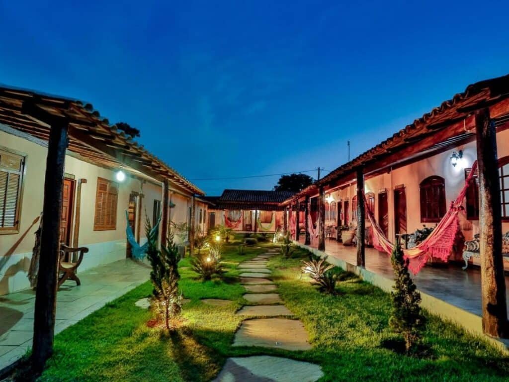 Um pátio com caminho de pedra, cercado por uma construção térrea com redes penduradas nas varandas, iluminado ao anoitecer. Esse é o Quatro Estações Pesqueiro e Hotel Fazenda, um dos hotéis fazenda em Minas Gerais.