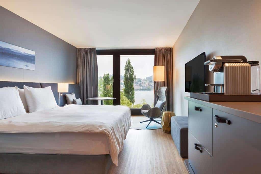 Foto do Radisson Blu Hotel, para representar o post de hotéis em Lucerna. A cama box de casal está encostada na parede à esquerda, e temos uma visão lateral dela. Na sua frente há uma cômoda com máquina de café e uma TV fixa na parede. No fundo do quarto há uma janela que vai do teto até o chão com vista, um abajur e uma poltrona no canto direito da parede.