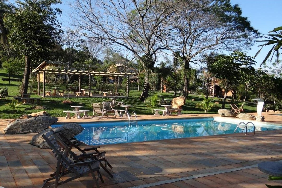 Piscina ao ar livre cercada por pátio de pedra, cadeiras de madeira e gramado verde com árvores e uma pérgola de madeira aberta ao fundo no Rancho Cipo Pousada, um dos hotéis fazenda em Minas Gerais.