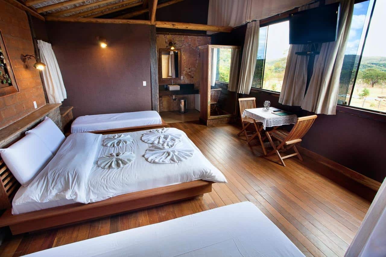 Um quarto de cabana de madeira no Racho Cipo Pousada com duas camas, uma mesa e cadeiras perto da janela, uma TV na parede e uma porta de banheiro nos fundos.