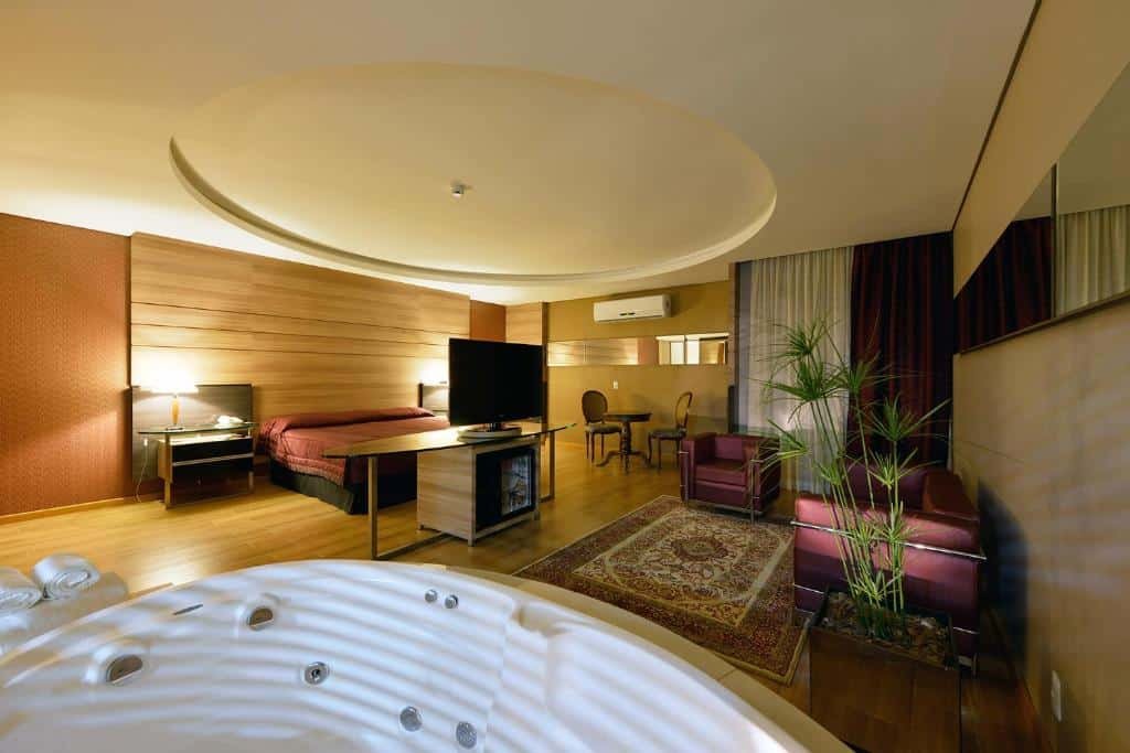 Quarto do Recanto Cataratas com uma banheira de hidromassagem e do lado um sofá, uma poltrona, um tapete no chão e uma tv. Ao fundo tem a cama de casal e uma mesinha do lado.