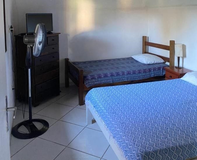 Quarto do Recanto Primavera. Uma cama de casal e uma de solteiro estão lado a lado e de frente para uma cômoda com uma tv e um ventilador.
