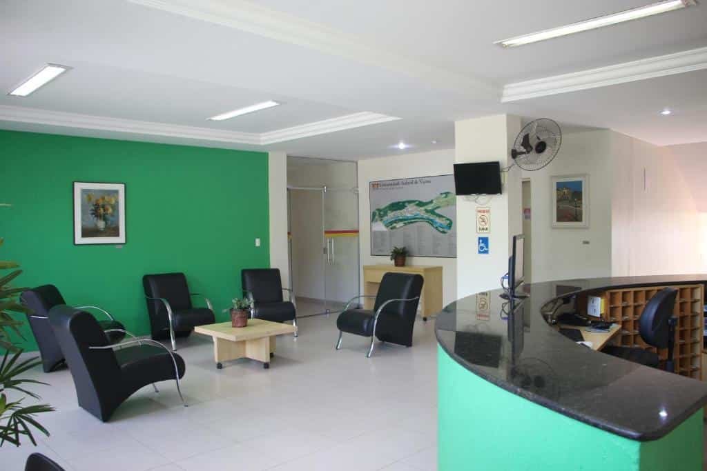 Recepção do Hotel do CEE. No lado direito há o balcão da recepção e no lado esquerdo estão cinco poltronas com uma mesa de centro e uma porta de vidro ao fundo.