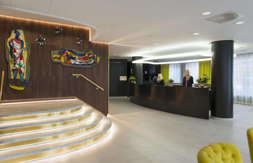 Recepção do Thon Hotel Rosenkrantz Oslo com balcão de atendimento em tom marrom com duas atendentes loiras e com blazer preto.